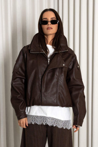 A model wearing a dark brown faux-leather moto jacket with a high collar, styled over a white top and pinstriped trousers, set against a neutral beige curtain background.