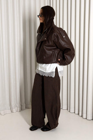 A person wearing a dark brown leather moto jacket, brown pinstripe wide-leg trousers, a white shirt with lace trim, and black shoes, standing in front of a white pleated curtain backdrop.