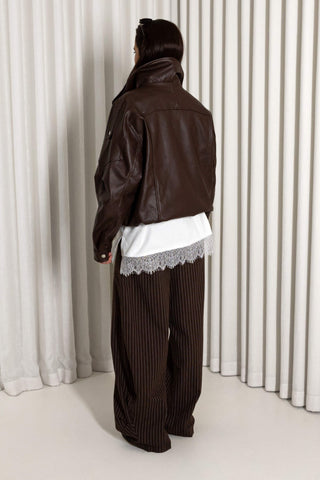 A person shown from the back wearing a dark brown leather jacket over a white lace-trimmed shirt and dark brown pinstriped trousers, standing in front of white pleated curtains.