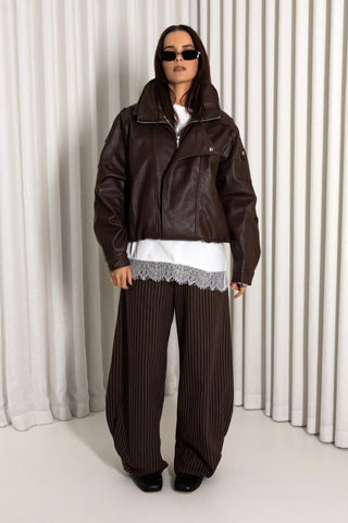 A model wearing a dark brown faux-leather moto jacket, matching dark brown pinstripe wide-leg trousers, a white t-shirt with a lace hem, and black sunglasses, standing in front of a white pleated curtain backdrop.