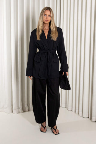 A woman modeling a black belted wrap jacket and matching wide-leg trousers, standing against a white pleated curtain backdrop.