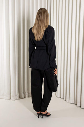 A woman modeling a black belted wrap jacket and matching wide-leg trousers, standing against a white pleated curtain backdrop.