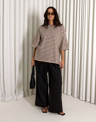 Woman wearing a striped shirt and black pants holding a black bag against a white curtain background High waisted black straight leg pants