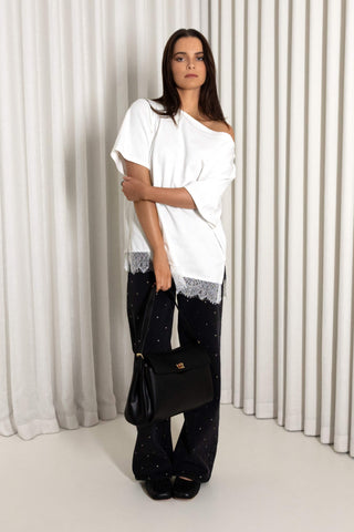 A woman stands in front of white pleated curtains wearing a white off-the-shoulder top with lace trim, black wide-leg pants with small white dots, and carrying a black handbag.