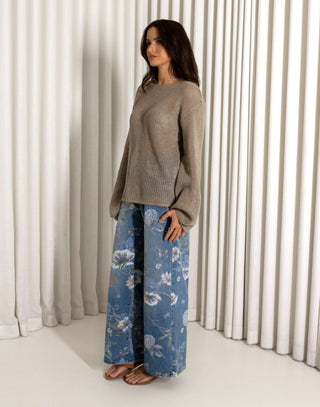 Woman wearing a beige sweater and blue floral jeans standing in front of white curtains. Beige open weave sheer knit long sleeve top