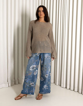 Woman wearing a beige sweater and blue floral jeans standing in front of white curtains. Beige open weave sheer knit long sleeve top