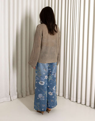 Woman wearing a beige sweater and blue floral jeans standing in front of white curtains. Beige open weave sheer knit long sleeve top