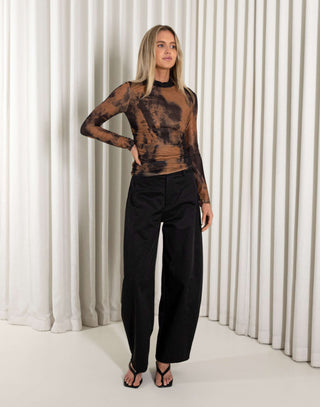 Woman wearing a black and brown tie-dye top with high-waisted black pants against a white curtain background.