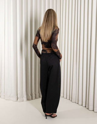 Woman wearing a black and brown tie-dye top with high-waisted black pants against a white curtain background.