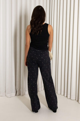 A woman viewed from behind wearing black wide-leg pants with a small white star pattern, a black sleeveless top, and holding a small cream clutch bag, set against a background of white vertical drapes.