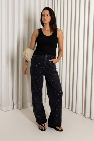 A woman stands in front of white pleated curtains wearing dark wide-leg jeans with a small white dot pattern, a black tank top, and black sandals, while holding a cream clutch.