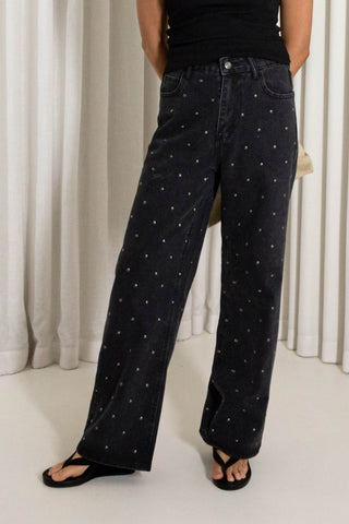 A person wearing dark washed wide-leg jeans with small white embroidered dots, styled with a black top and black sandals in front of white curtains.