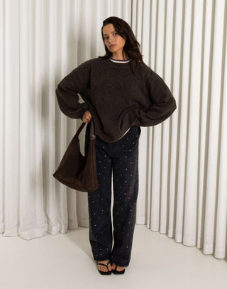 Woman wearing a brown oversized knit and  black straight leg stud jeans standing against a white curtain.