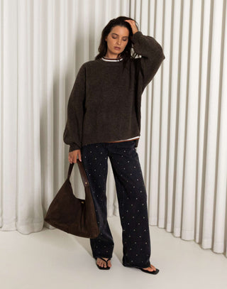 Woman wearing a brown oversized knit and  black straight leg stud jeans standing against a white curtain.