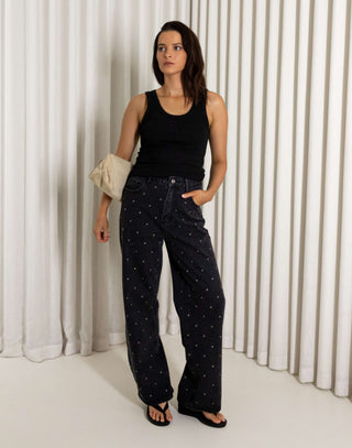 Woman wearing a black tank top and black straight leg stud jeans standing against a white curtain.