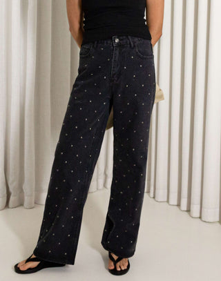 Woman wearing a black tank top and black straight leg stud jeans standing against a white curtain.