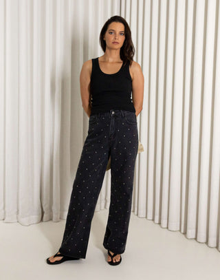 Woman wearing a black tank top and black straight leg stud jeans standing against a white curtain.