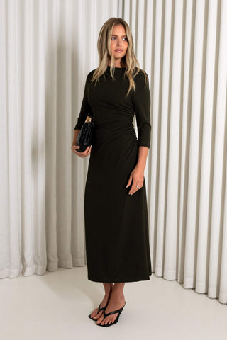 A woman modeling a olive midi dress with three-quarter sleeves, a high neckline, and a gathered waist detail, standing against a white pleated curtain backdrop.