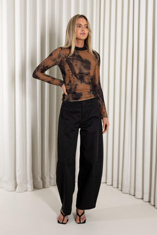 A woman modeling a sheer brown and black abstract-patterned long-sleeve top paired with black wide-leg trousers and black strappy heels, standing in front of a white pleated curtain backdrop.