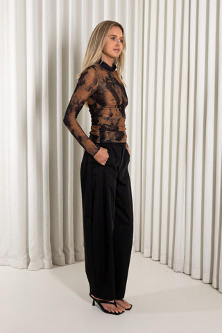 A woman modeling a sheer brown and black abstract-patterned long-sleeve top paired with black wide-leg trousers and black strappy heels, standing in front of a white pleated curtain backdrop.