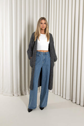 A woman modeling a long, open-front charcoal gray knit cardigan over a white cropped top and blue wide-leg jeans, standing against a white curtain backdrop.