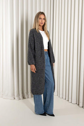 A woman modeling a long, open-front charcoal gray knit cardigan over a white cropped top and blue wide-leg jeans, standing against a white curtain backdrop.