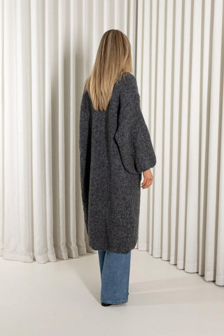A woman modeling a long, open-front charcoal gray knit cardigan over a white cropped top and blue wide-leg jeans, standing against a white curtain backdrop.