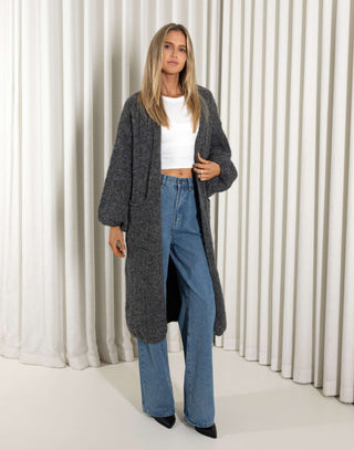 Woman wearing a long dark gray cardigan, white top, and blue jeans against a white curtain background.