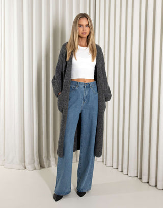 Woman wearing a long dark gray cardigan, white top, and blue jeans against a white curtain background.