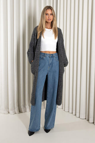 A woman stands in front of a white pleated curtain backdrop wearing a long dark gray open-front cardigan over a white cropped top and high-waisted wide-leg blue jeans.