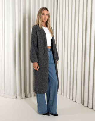Woman wearing a long dark gray cardigan, white top, and blue jeans against a white curtain background.