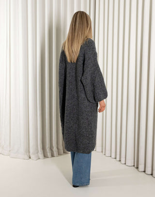 Woman wearing a long dark gray cardigan, white top, and blue jeans against a white curtain background.