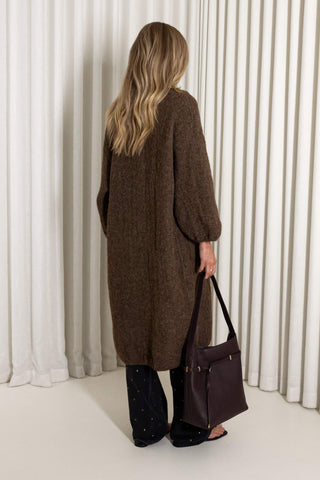 A woman shown from the back wearing a long brown knit cardigan, dark wide-leg pants, and holding a dark brown leather tote bag, standing in front of white pleated curtains.