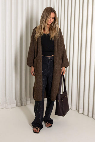 A woman stands in a studio wearing a long brown textured open-front cardigan over a black top and dark patterned pants, holding a dark brown tote bag. The background features white vertical drapes.