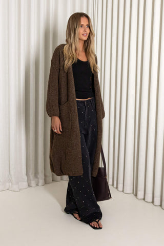 A woman stands in front of white pleated curtains wearing a long brown textured open-front coat over a black cropped top and dark patterned wide-leg pants, with a dark shoulder bag.