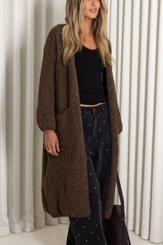 A woman wearing a long brown open-front cardigan over a black top and dark patterned pants, standing in front of white vertical drapes.