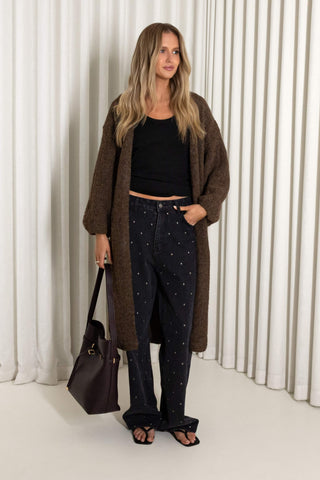 A woman stands in a studio wearing a brown open-front cardigan over a black cropped top, dark patterned pants, and black sandals, while holding a dark purple handbag. She is positioned in front of a white pleated curtain backdrop.