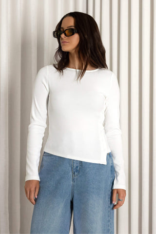 A woman wearing a fitted white long-sleeve top and light blue jeans, standing in front of a cream-colored pleated curtain backdrop.