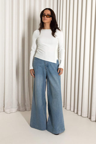 A woman modeling a white long-sleeve top and wide-leg blue denim jeans, standing in front of a beige pleated curtain backdrop.