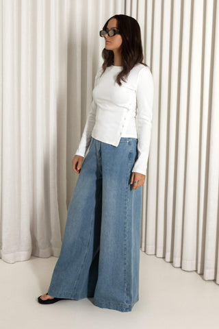 A woman wearing a white long-sleeve top and wide-leg blue denim jeans, standing in front of a white pleated curtain backdrop.