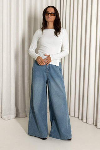 A woman modeling a white long-sleeve top and wide-leg blue denim jeans against a background of vertical white drapes.