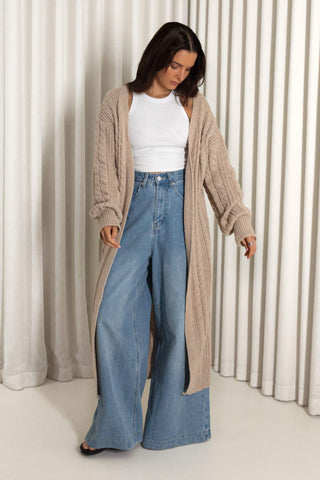A woman wearing a long beige cable-knit cardigan over a white ribbed top and high-waisted wide-leg blue jeans, standing in front of a cream-colored pleated curtain backdrop.