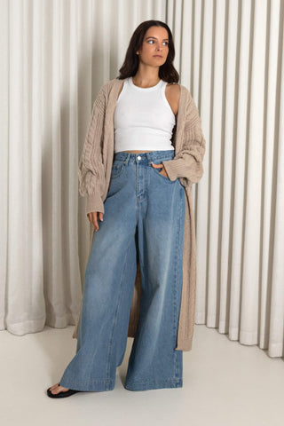 A woman stands in front of cream pleated curtains wearing a beige knit cardigan over a white ribbed tank top and light blue wide-leg denim jeans.