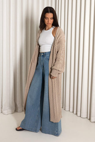 A woman stands indoors wearing a long beige cable-knit cardigan over a white tank top and wide-leg blue jeans, with black sandals, in front of cream pleated curtains.