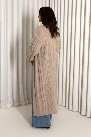 A woman shown from the back wearing a long, chunky cable-knit cardigan in a light beige color, layered over blue jeans, standing in front of white vertical drapes.