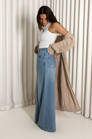 A woman modeling a pair of wide-leg blue denim jeans, styled with a white sleeveless top and a beige cable-knit cardigan draped over her shoulders, set against a background of cream-colored curtains.