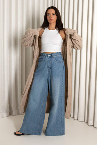 A woman modeling a pair of wide-leg light blue denim jeans, styled with a white ribbed tank top and a beige open-front cardigan, standing in front of a cream-colored pleated curtain backdrop.
