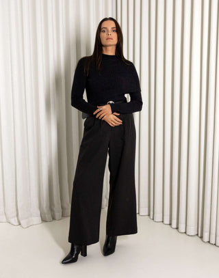 Woman wearing a black turtleneck and black pants with a belt against a white curtain background