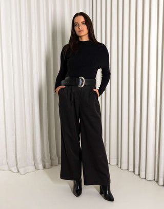 Woman wearing a black turtleneck and black pants with a belt against a white curtain background