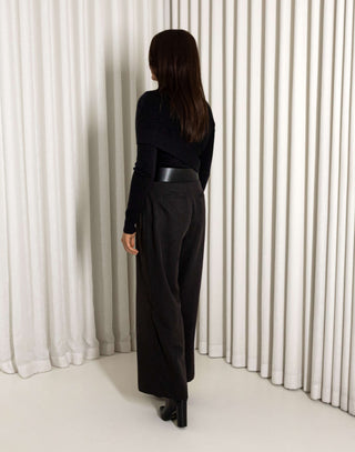 Woman wearing a black turtleneck and black pants with a belt against a white curtain background